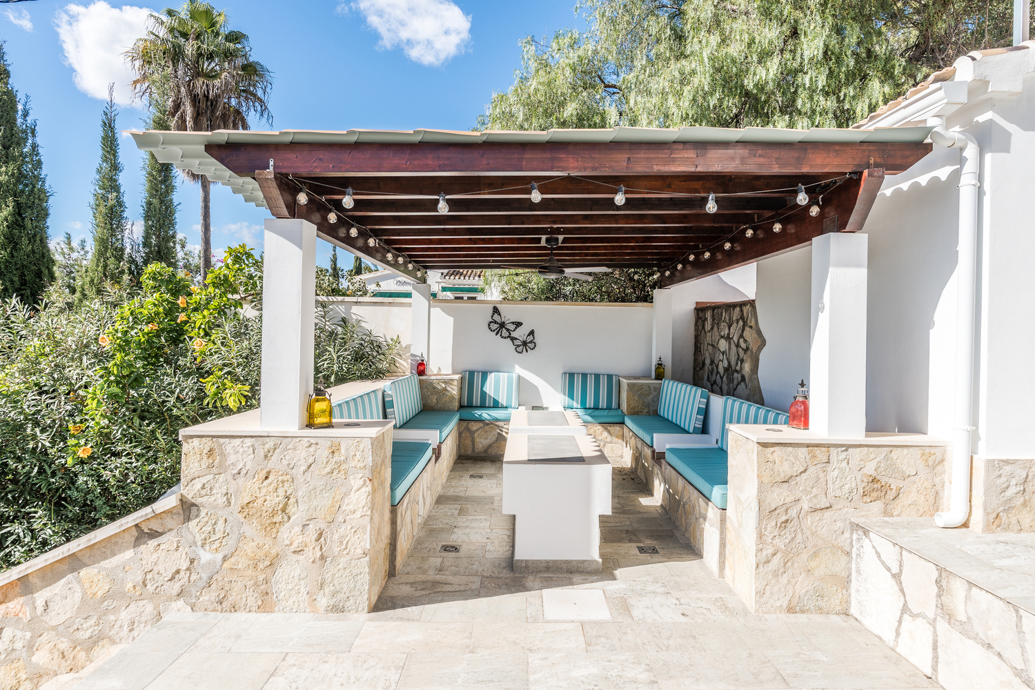Überdachte Außenlounge mit Holzpergola, Lichterkette und integrierten Steinbänken mit türkisfarben gestreiften Polstern und Deckenventilator.