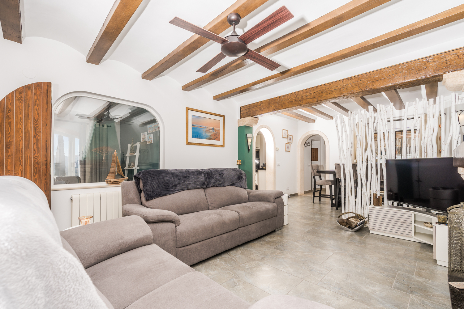 Wohnbereich mit rustikalen Holzbalken, Deckenventilator und Fliesenboden. Ein dekorativer Raumteiler aus Ästen trennt die Sitzecke vom angrenzenden Esszimmer.