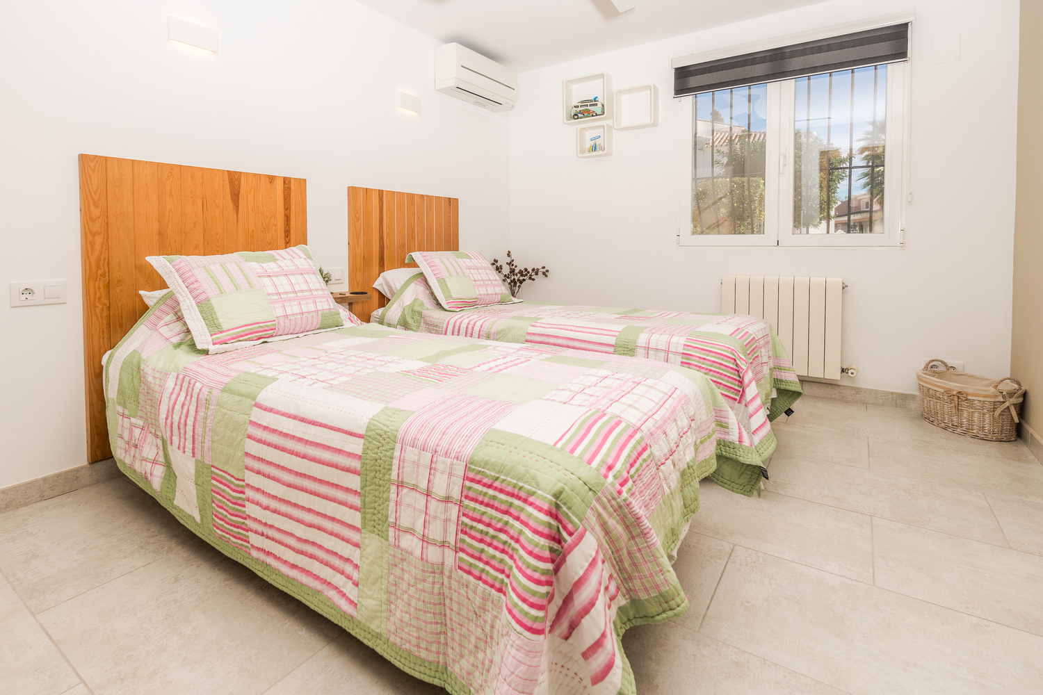 Zweibettzimmer mit hölzernen Kopfteilen, hellem Fliesenboden und Klimaanlage. Das Zimmer bietet ein Fenster mit Sicherheitsgittern und einen Heizkörper.