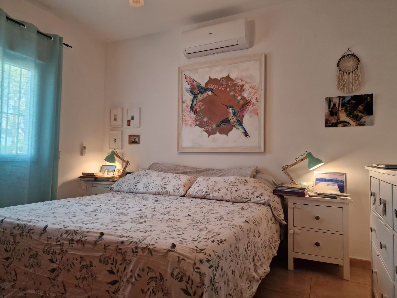 Helles Schlafzimmer mit Doppelbett, Haier-Klimaanlage, weißen Nachttischen mit grünen Leselampen und einem großen Kolibri-Wandbild über dem Bett.