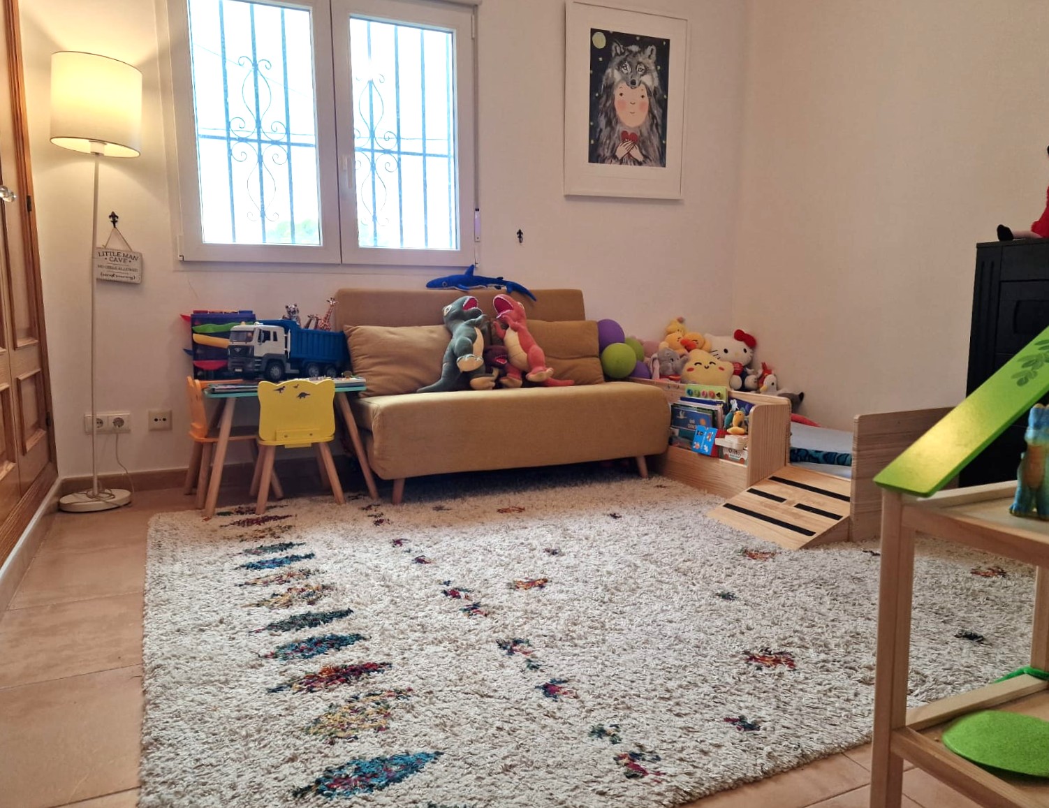 Kinderbereich mit strukturiertem Teppich, gelbem Sofa und kleinem Spieltisch. Der helle Raum verfügt über Fliesenboden und ein Fenster mit dekorativen Sicherheitsgittern.