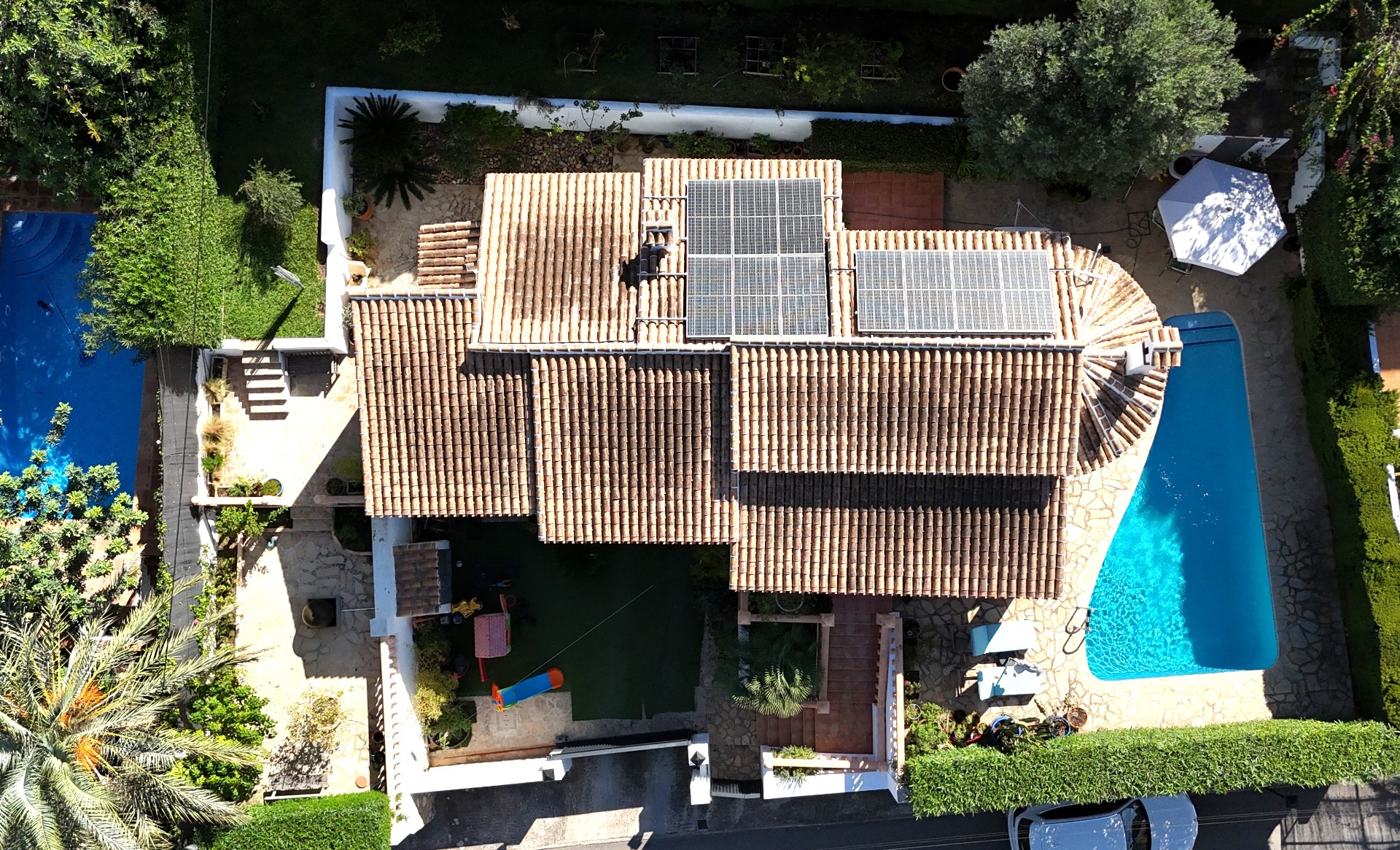 Luftbild der Villa mit Terrakotta-Ziegeldach und Solaranlage, privatem Pool und einem mediterranen Garten inklusive Kinderspielplatz und Steinpflaster.