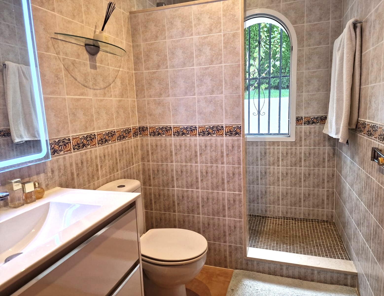 Modernes Badezimmer mit begehbarer Dusche, beigen Fliesen mit dekorativer Bordüre und einem Rundbogenfenster, das für natürliches Licht sorgt.