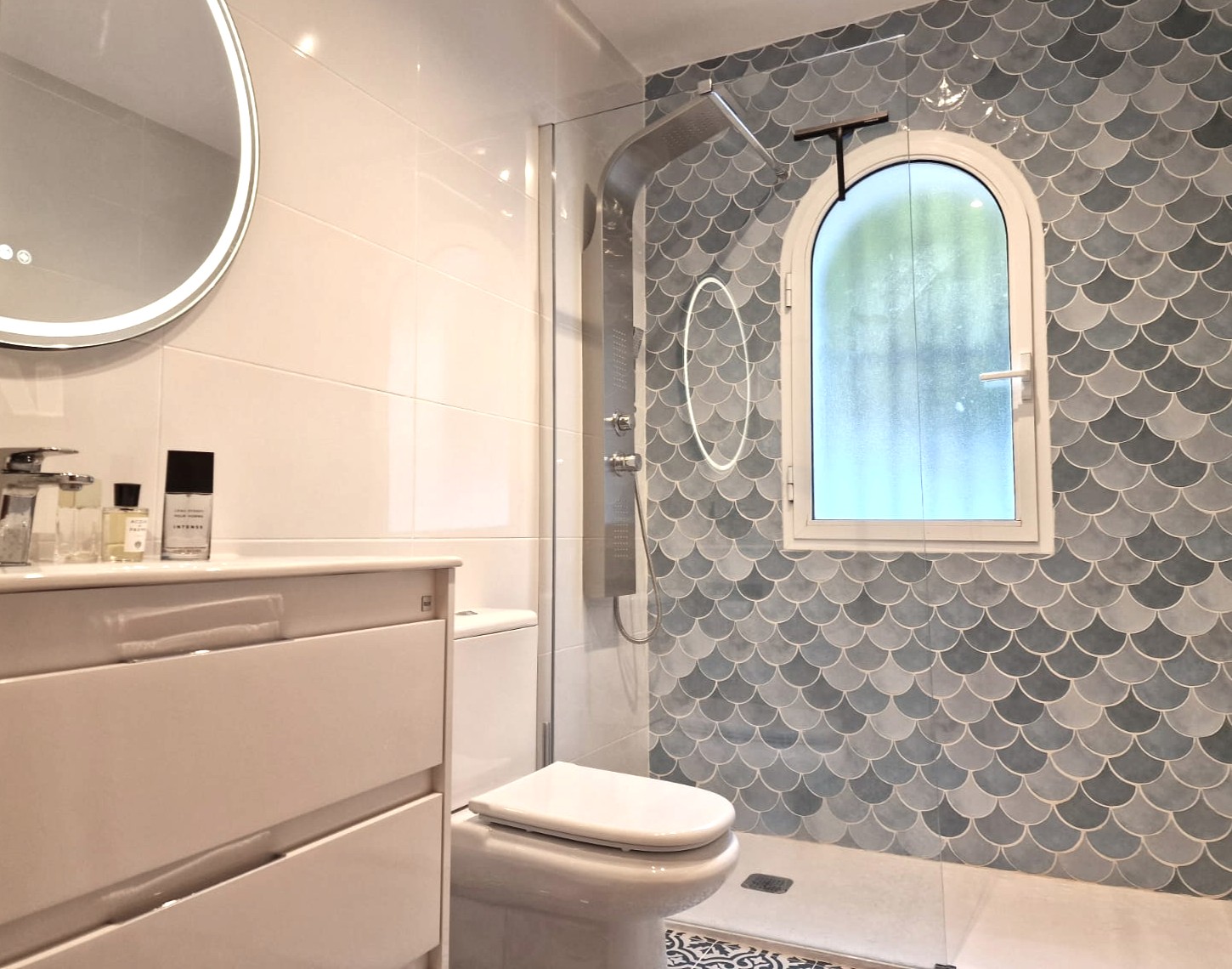 Modernes Badezimmer mit begehbarer Dusche, blauen Schuppenfliesen, Rundbogenfenster mit Milchglas, Edelstahl-Duschsäule und hinterleuchtetem Rundspiegel.