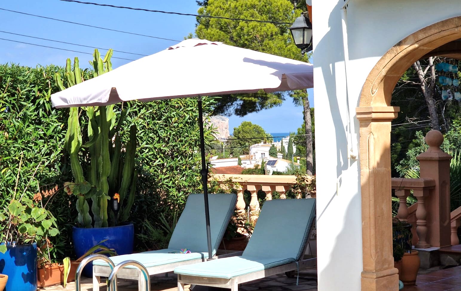 Terrassenbereich mit Sonnenliegen und Sonnenschirm neben einem Steinbogen, umgeben von mediterraner Flora mit Fernblick auf das Meer und das Cabo de San Antonio.