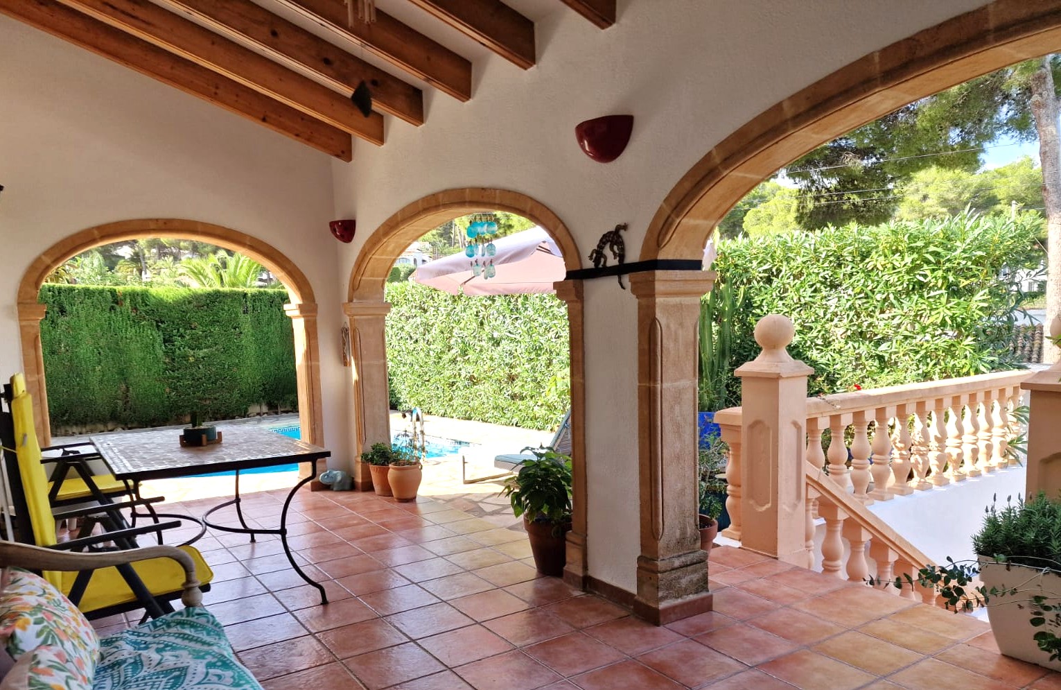 Überdachte Terrasse mit Bögen aus Tosca-Stein, sichtbaren Holzbalken und Terrakottaboden, mit Blick auf den privaten Pool und den Garten.