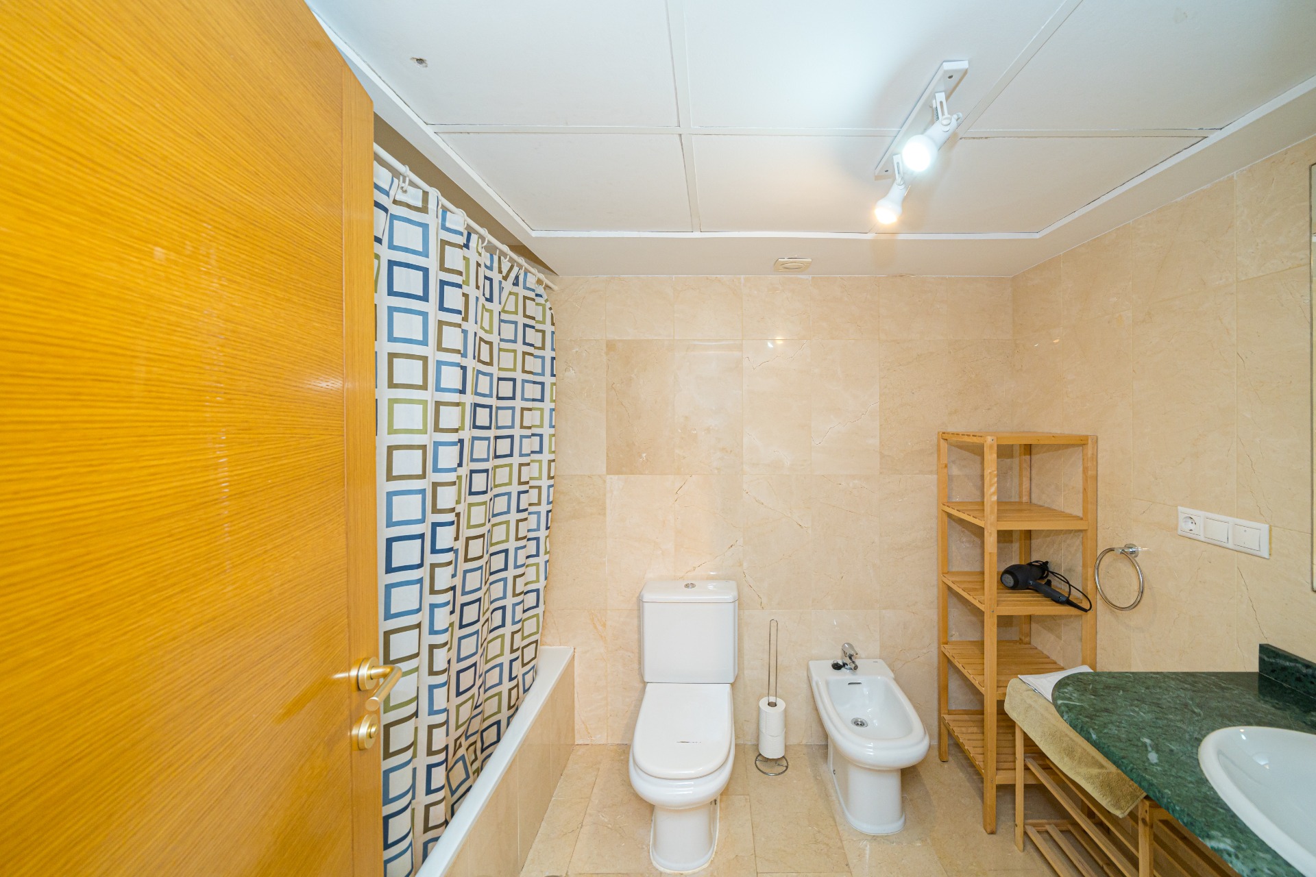 Badezimmer mit beigen Marmorfliesen, ausgestattet mit Badewanne, Bidet und einer Waschtischplatte aus grünem Marmor sowie praktischen Holzregalen.