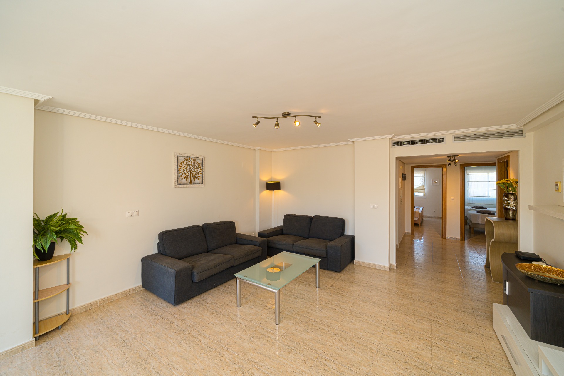 Großzügiger Wohnbereich mit dunkelgrauen Sofas, Glastisch und poliertem Fliesenboden. Der offene Grundriss führt direkt zum Flur der Schlafräume.