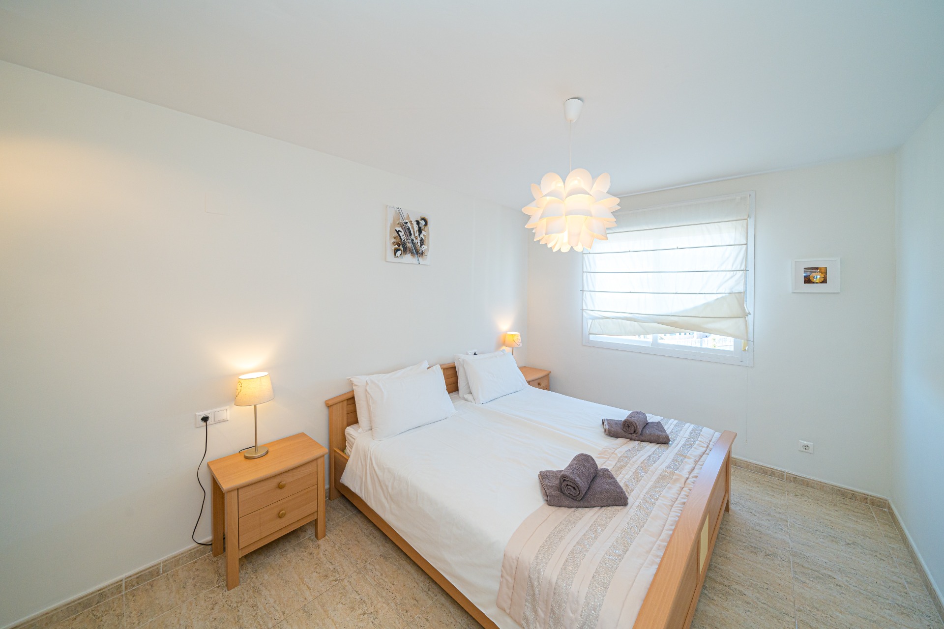 Helles Schlafzimmer mit hellen Holzmöbeln, Doppelbett und passenden Nachttischen. Natürliches Licht fällt durch das Fenster mit weißem Rollo auf den Fliesenboden.