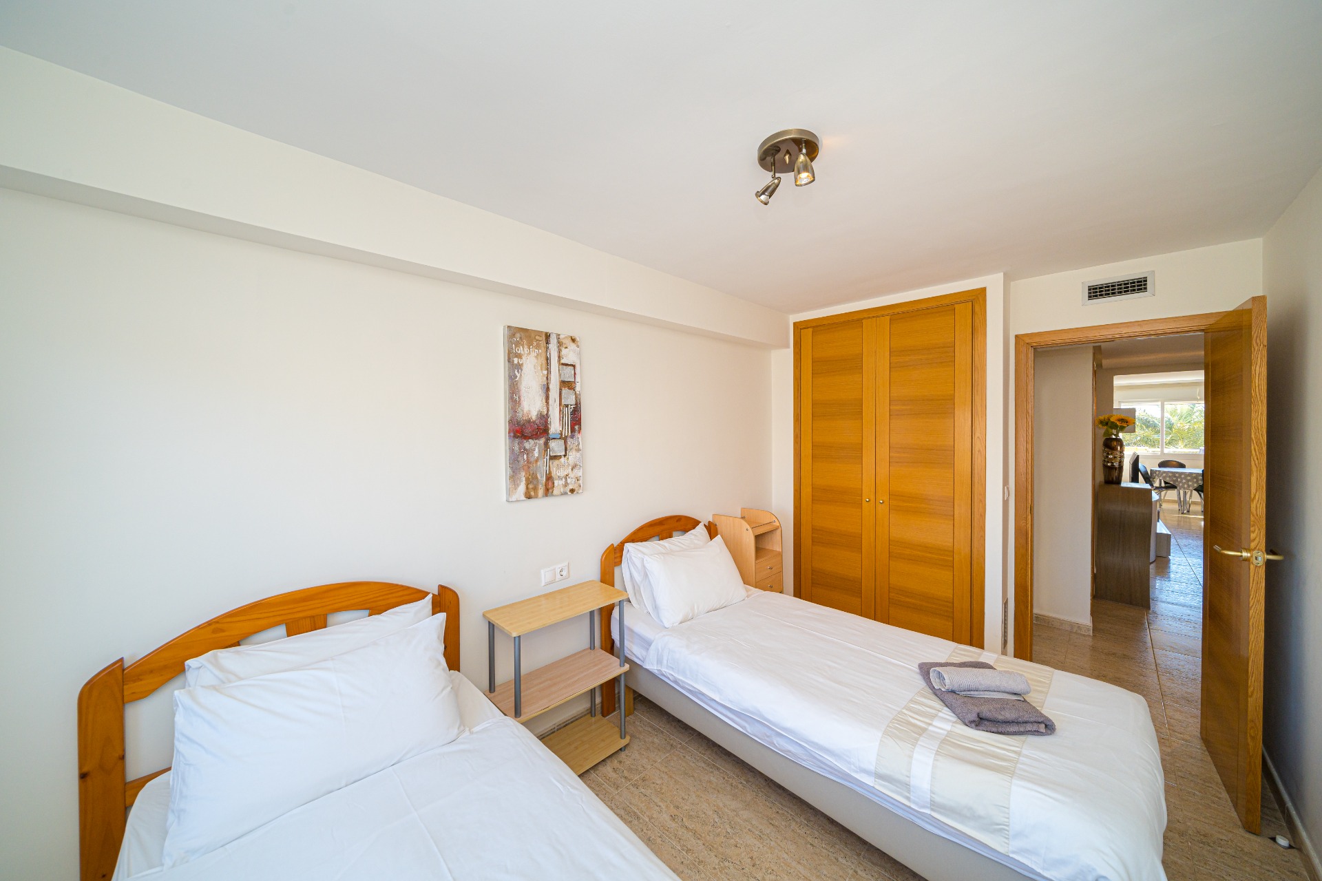 Helles Schlafzimmer mit zwei Einzelbetten, hölzernen Kopfteilen, einem Einbauschrank in Eichenoptik und Fliesenboden. Eine offene Tür führt zum Wohnbereich.
