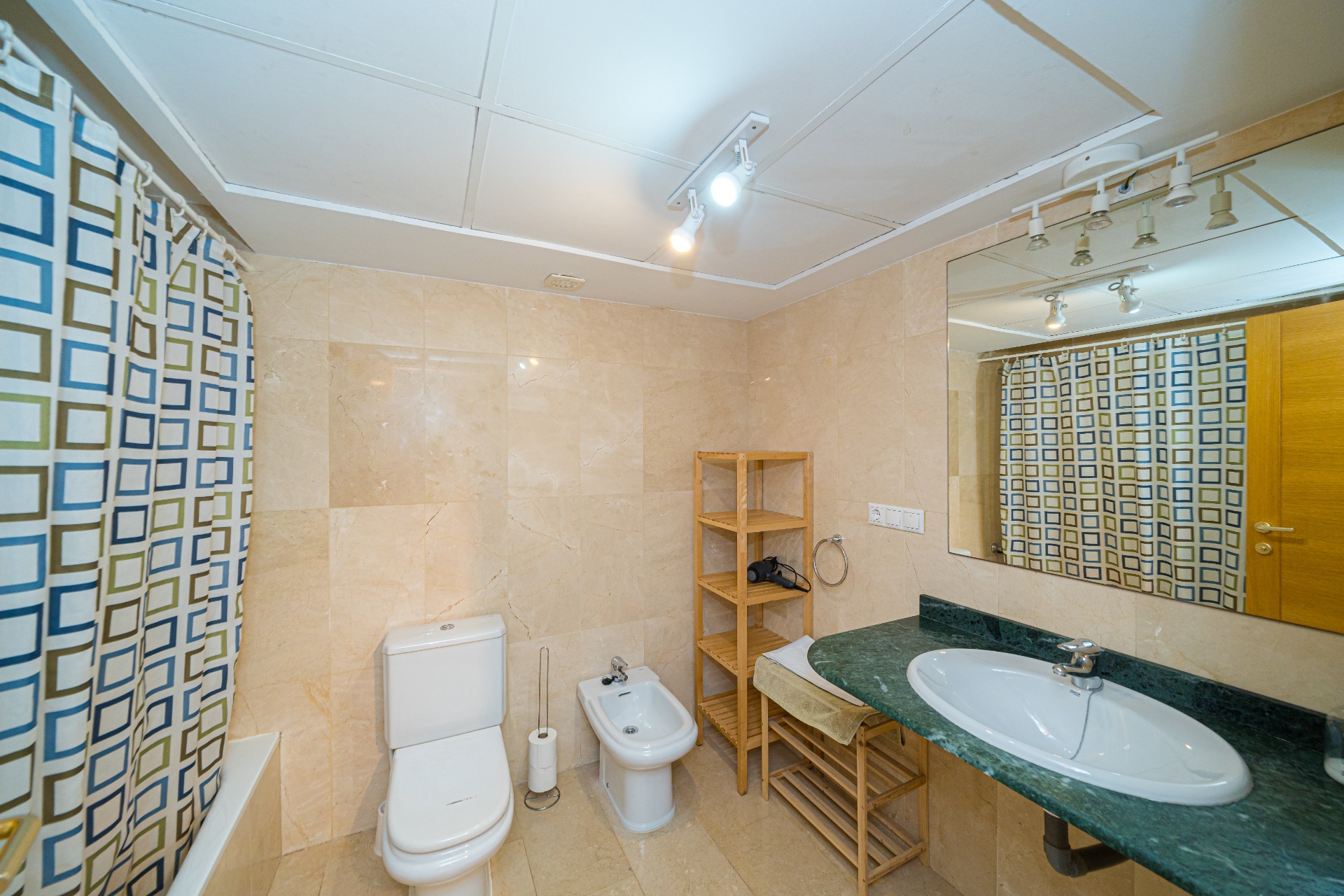 Modernes Badezimmer mit cremefarbenen Marmorfliesen, grünem Marmorwaschtisch und Bidet. Ausgestattet mit Holzregalen, einer Badewanne und Deckenstrahlern.