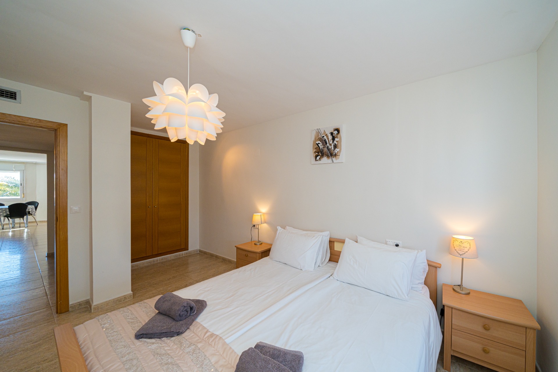 Schlafzimmer mit Doppelbett, Einbauschränken aus Holz und einer dekorativen Hängeleuchte. Der Blick durch die Tür führt in den hellen Essbereich der Wohnung.