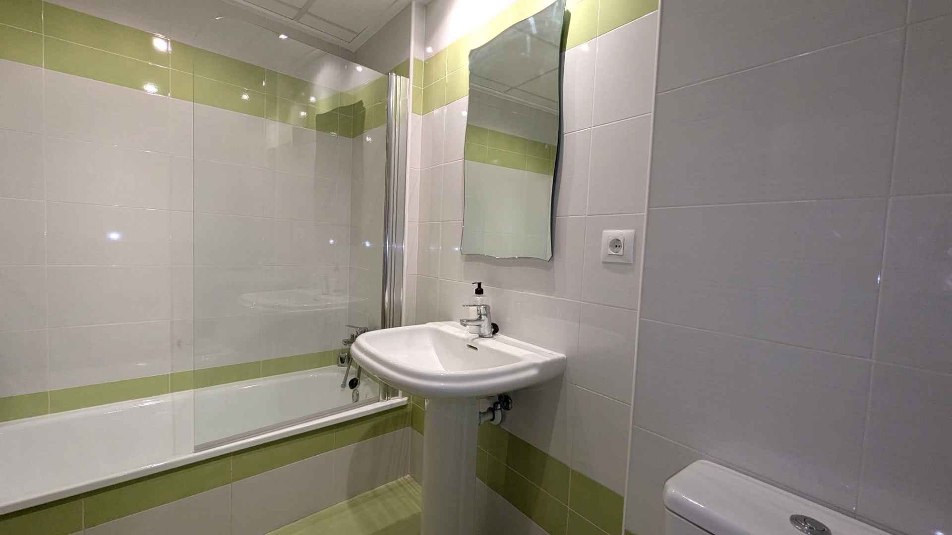 Badezimmer mit Badewanne, Glastrennwand und Standwaschbecken, gestaltet mit weiß-grünen Fliesen und einem Spiegel mit Wellenschnitt.