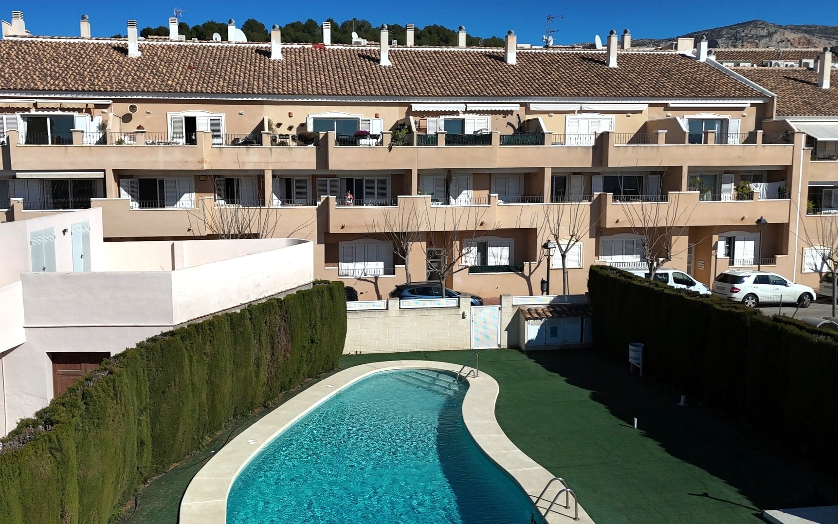 Blick auf den gemeinschaftlichen nierenförmigen Pool, umgeben von Kunstrasen und hohen Hecken, vor der Fassade des Wohnkomplexes mit weißen Fensterläden.