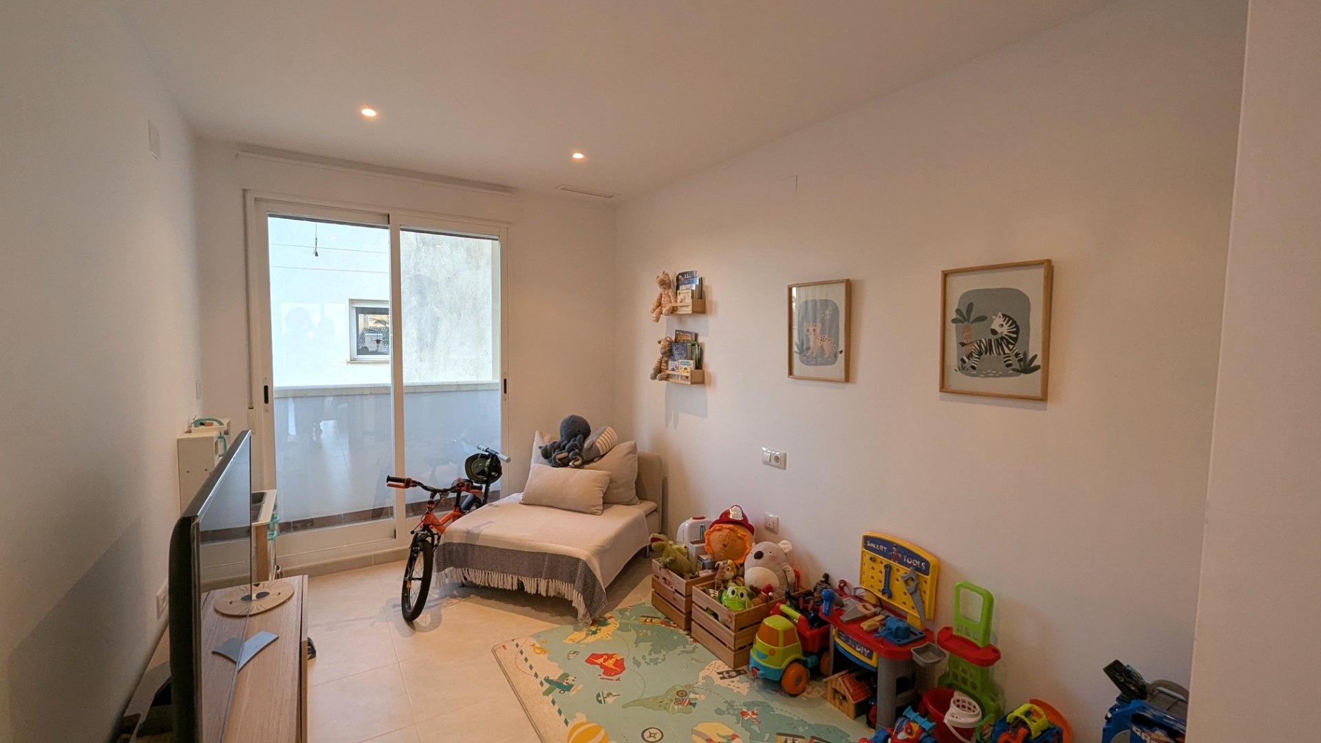 Helles Schlafzimmer, derzeit als Spielzimmer genutzt, mit Deckeneinbaustrahlern, hellen Fliesenböden und Glasschiebetüren zum privaten Balkon.