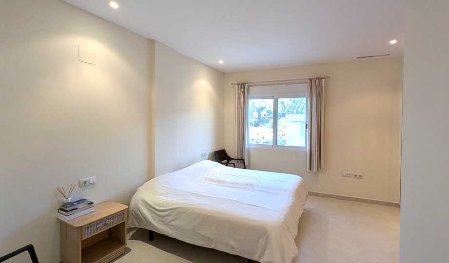 Helles Schlafzimmer in neutralen Tönen mit Deckeneinbaustrahlern und Fenster mit Rollläden. Ausgestattet mit Fliesenboden und einem Nachttisch aus Holz.