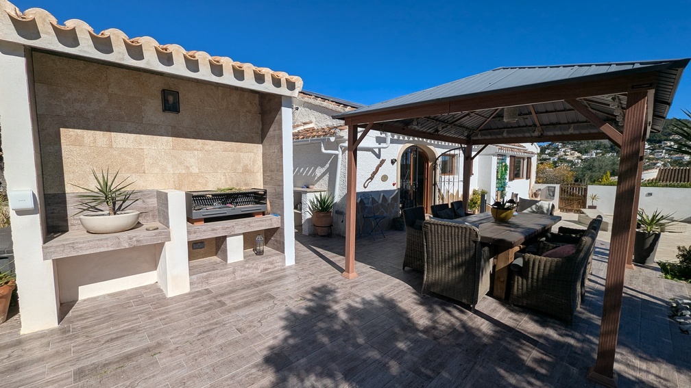 Außen-Essbereich mit gemauertem Grillplatz aus Naturstein und einer großen Pergola über einer Sitzgruppe mit Korbstühlen auf Fliesen in Holzoptik.