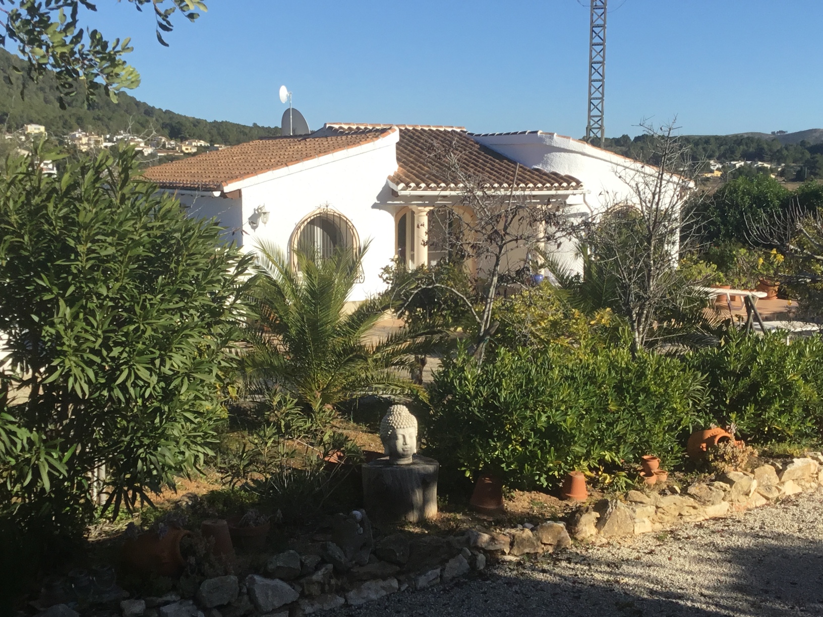 Außenansicht der weiß getünchten mediterranen Villa mit Rundbogenfenstern, Terrakotta-Dachziegeln und angelegtem Garten mit Palmen und einer Buddha-Statue.