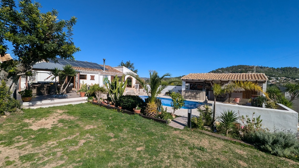 Außenansicht des Haupthauses mit Solarmodulen, privatem Pool und überdachter Außenküche in einem mediterran gestalteten Garten mit Weitblick.