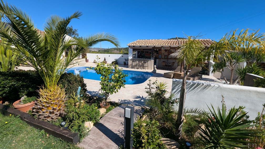 Außenpoolbereich mit Steinterrasse, mediterraner Gartenanlage mit Palmen und einer überdachten Sommerküche mit Bar und Barhockern.