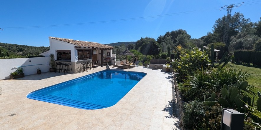 Außenpoolbereich mit Steinterrasse, voll ausgestatteter Sommerküche mit Bar und mediterran bepflanztem Garten mit Blick auf die umliegende Landschaft.