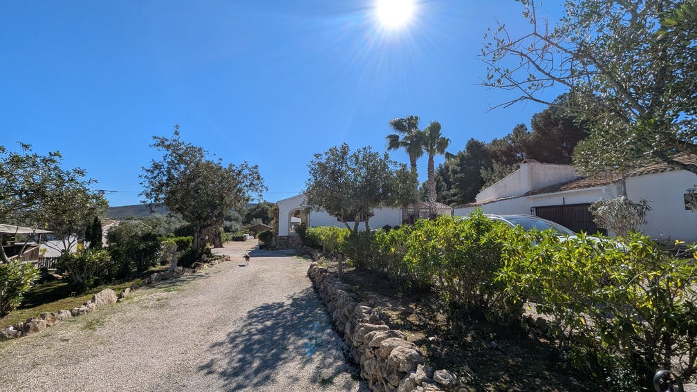 Breite Kiesauffahrt mit traditionellen Steinmauern, die zum Haupthaus im Finca-Stil führt, umgeben von mediterranen Bäumen und Palmen unter blauem Himmel.