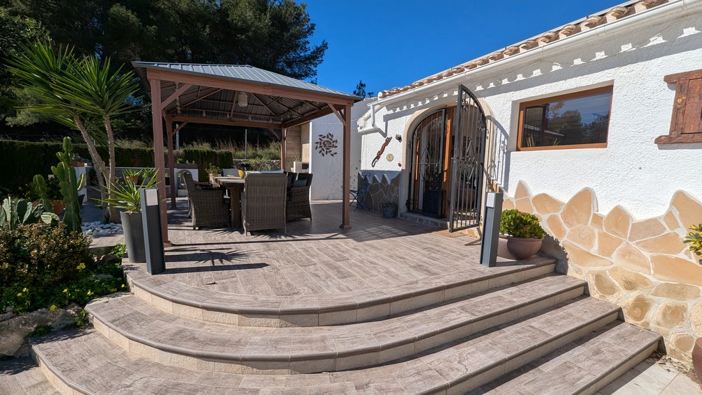 Erhöhte Terrasse mit Fliesen in Steinoptik und geschwungenen Stufen, die zu einem schattigen Essbereich unter einer Holzpergola führen, ergänzt durch eine weiße Fassade mit Naturstein.