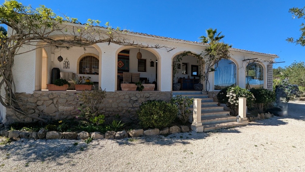 Finca-Fassade mit traditioneller bogenförmiger Naya, Natursteinsockel und Außentreppe, ergänzt durch mediterrane Kletterpflanzen.