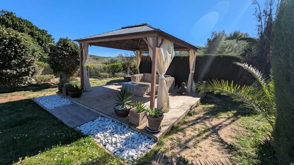 Gartenpavillon aus Holz auf einer Echtholzterrasse mit Rattan-Sitzgruppe, hellen Vorhängen und dekorativen weißen Kieselsteinen in einer gepflegten Außenanlage.
