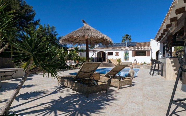 Geflieste Terrasse mit privatem Pool, Korbliegen unter einem Reet-Sonnenschirm und Außenbar. Das Haupthaus verfügt über Solarmodule auf dem Ziegeldach.