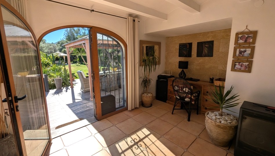Heller Arbeitsbereich mit Terrakottaboden, Natursteinwand und bogenförmigen Holztüren, die auf eine sonnige Gartenterrasse mit Pergola führen.