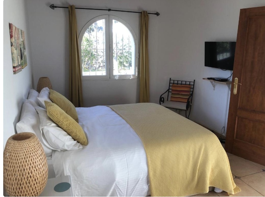 Helles Schlafzimmer mit Rundbogenfenster, Terrakottaboden und wandmontiertem Fernseher. Die Einrichtung besticht durch senfgelbe Akzente und geflochtene Lampen.