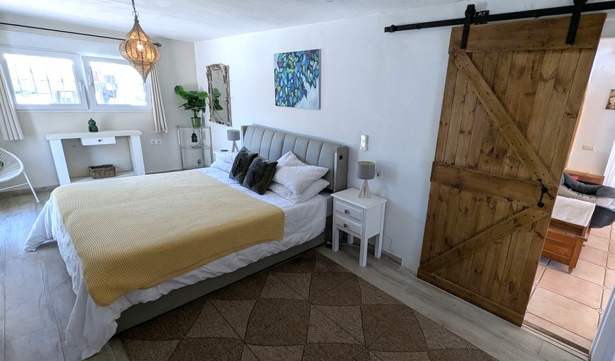 Helles Schlafzimmer mit rustikaler Holzschiebetür, gepolstertem Kopfteil und dekorativer marokkanischer Hängelampe auf Boden in Holzoptik.