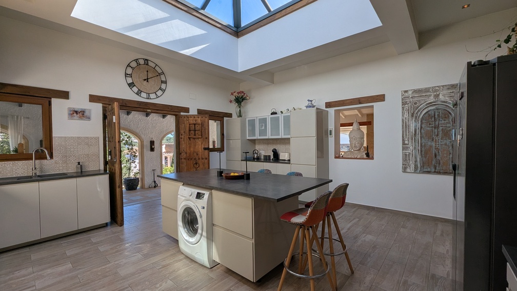 Moderne Küche mit zentraler Insel und großem Glas-Oberlicht. Traditionelle Holztüren und ein Durchblick zur Naya betonen den mediterranen Charakter des Hauses.
