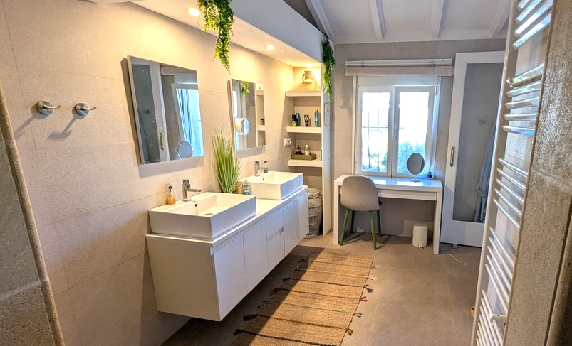 Modernes Badezimmer mit Doppelwaschtisch, schwebenden Schränken und Einbauregalen. Ein Schminktisch am Fenster und weiße Deckenbalken sorgen für ein helles Ambiente.