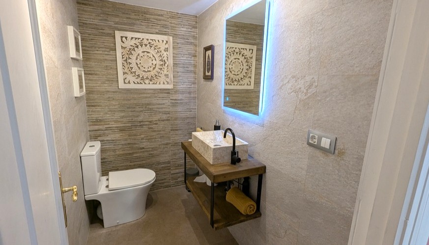 Modernes Gäste-WC mit Steinwaschbecken auf einem schwebenden Holzregal, hinterleuchtetem LED-Spiegel und strukturierten Fliesen in Natursteinoptik.