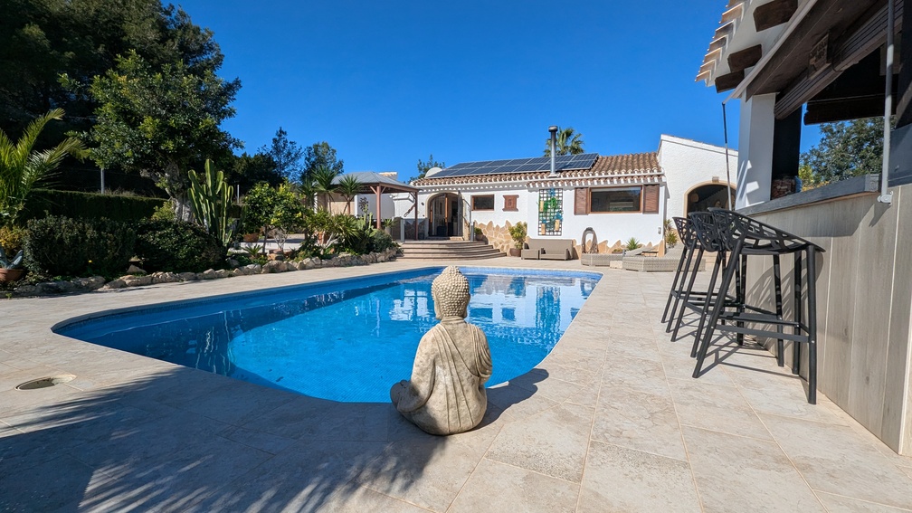 Privater Poolbereich mit Natursteinfliesen, Buddha-Statue, Außenbar mit Hockern und Villa mit installierter Photovoltaikanlage auf dem Dach.