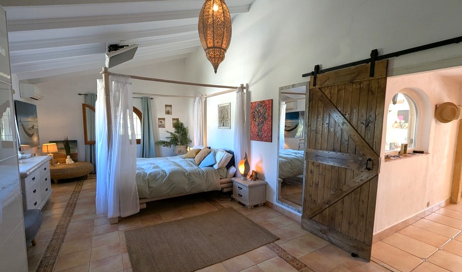 Schlafzimmer mit Himmelbett, weißen Balkendecken und rustikaler Scheunentür. Terrakottafliesen und eine marokkanische Lampe betonen den mediterranen Stil.