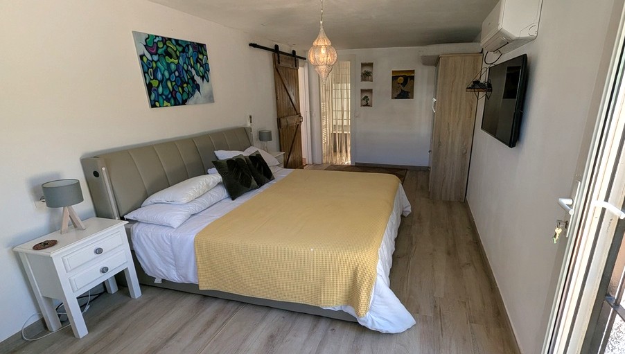 Schlafzimmer mit Holzboden, rustikaler Schiebetür im Scheunenstil und Hängelaterne. Ausgestattet mit Klimaanlage, Wand-TV und einem Bett mit grauem Polsterkopfteil.