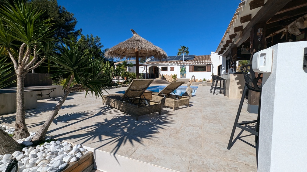 Sonnige Fliesenterrasse mit Korbliegen, Strohschirm und Pool, im Hintergrund das Haupthaus mit sichtbarer Photovoltaikanlage auf dem Ziegeldach.