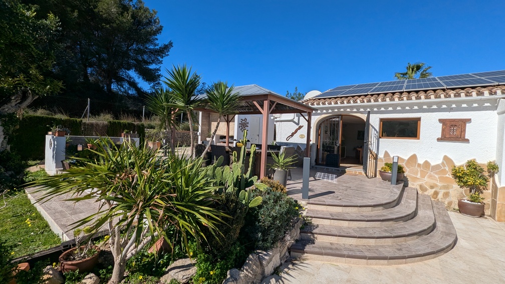 Terrasse mit geschwungenen Steinstufen, Holzpavillon für Mahlzeiten im Freien und weiße Fassade mit Photovoltaikanlage und mediterraner Bepflanzung.
