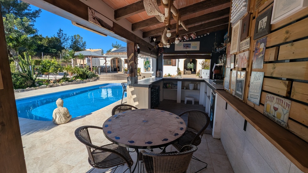Überdachte Außenküche mit Bar, Holzbalkendecke und Mosaiktisch mit Blick auf den privaten Pool und die mediterrane Gartenanlage.