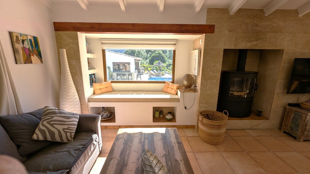 Wohnbereich mit integrierter Fensterbank und Poolblick, modernem Kaminofen in einer Tosca-Steinwand und traditionellen Holzbalkendecken.