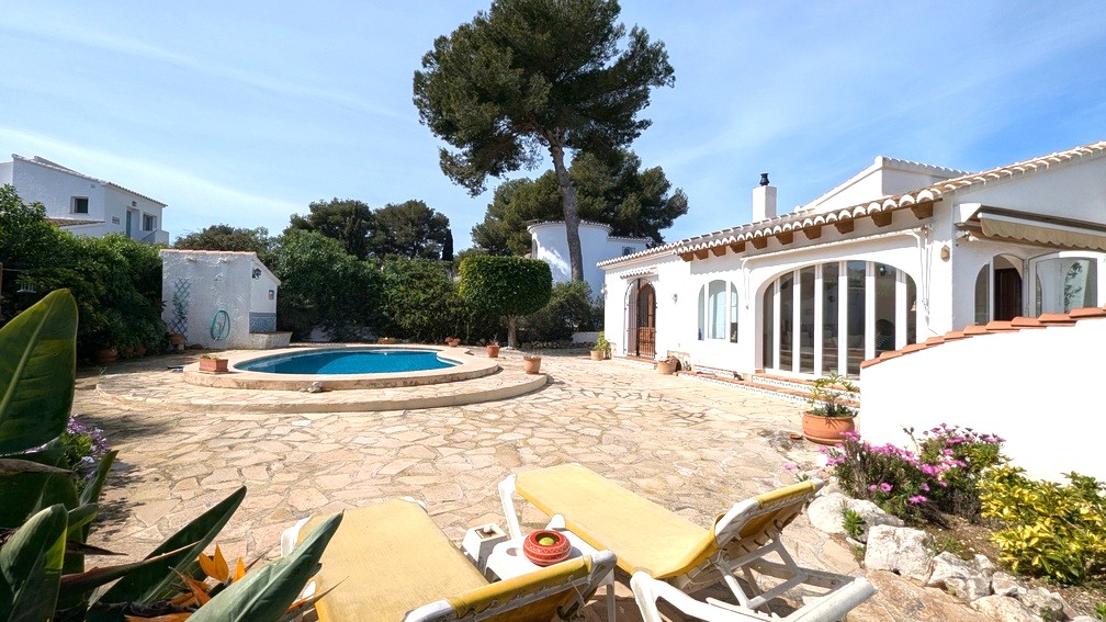 Außenansicht der mediterranen Villa mit Pool, Natursteinterrasse und weißer Fassade mit sichtbaren Holzbalken und großen bogenförmigen Glastüren.