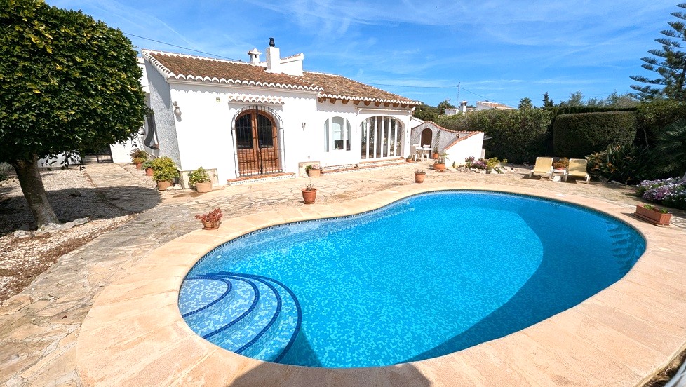 Außenansicht des nierenförmigen Pools mit blauen Mosaikfliesen, umgeben von einer Steinterrasse und der traditionellen weißen Villa mit Terrakottadach.