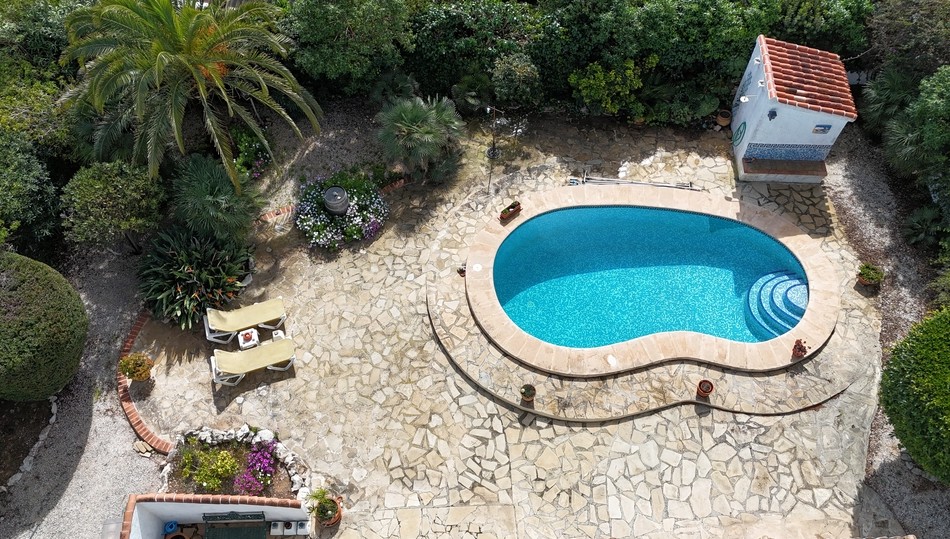 Erhöhte Ansicht eines nierenförmigen Pools mit blauen Mosaikfliesen, umgeben von einer Natursteinterrasse und einem eingewachsenen mediterranen Garten mit Palmen.