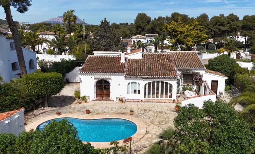 Erhöhte Außenansicht der Villa mit nierenförmigem Pool, Natursteinterrasse und traditionellem Ziegeldach mit Blick auf den fernen Berg Montgó.