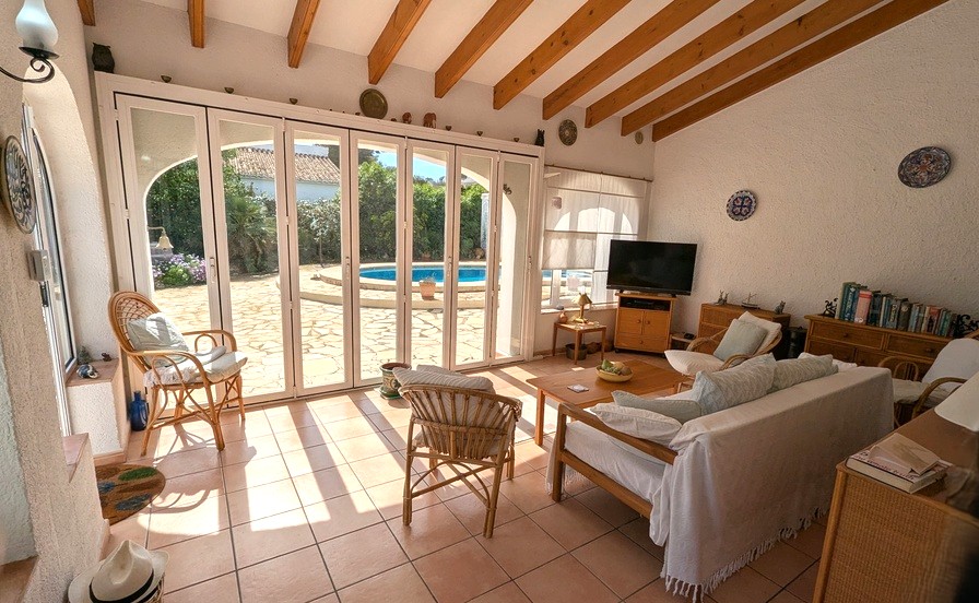 Helles Wohnzimmer mit sichtbaren Holzbalken, Terrakottaboden und großen Glasfalttüren, die direkt auf die Poolterrasse führen.
