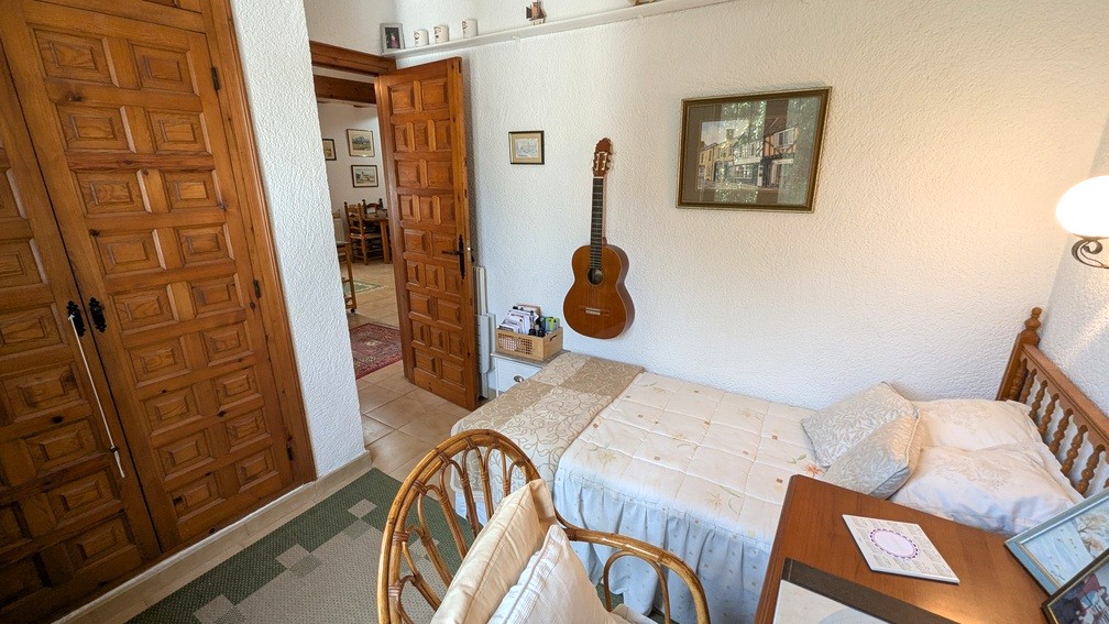 Schlafzimmer mit Einzelbett, Einbauschränken aus Holz mit traditionellen Paneelen und weißen Strukturwänden. Die offene Tür führt zum Flur und Wohnbereich.