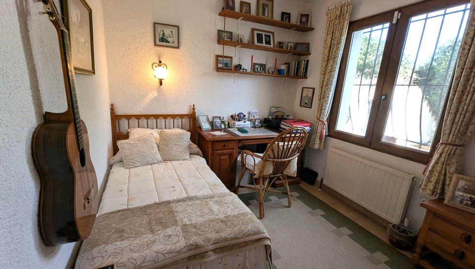 Schlafzimmer mit Einzelholzbett, klassischem Schreibtisch, Korbstuhl und Wandregalen. Inklusive Heizkörper und Fenster mit dekorativen Sicherheitsgittern.